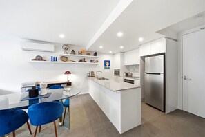 A modern kitchen with white cabinetry, a stainless steel refrigerator, a glass dining table with blue chairs, and a built-in island with a sink and microwave. - Stylish 1-Bed Retreat in Braddon (Braddon)