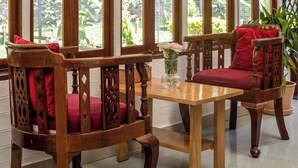 Una habitación con una mesa de madera y dos sillas con cojines rojos, ventanas amplias que dan a zonas verdes, y suelo de baldosas.