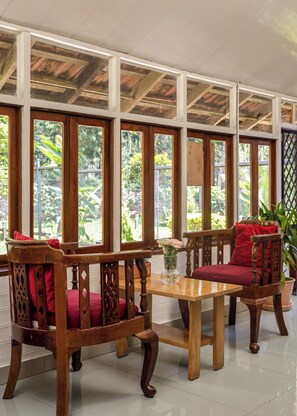 A room with a wooden table and two chairs with red cushions, large windows with a view of greenery, and a tiled floor.