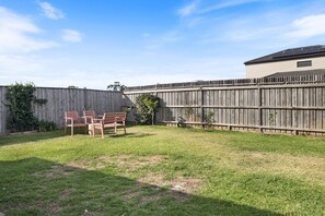 A backyard with a wooden fence, a patio with chairs, and a building with a sloped roof. - Garden Retreat 4-Bed House in Tarneit (Tarneit)