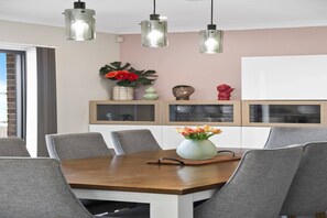 A dining area with a wooden table, grey chairs, pendant lights, and decorative items on shelves. - Garden Retreat 4-Bed House in Tarneit (Tarneit)