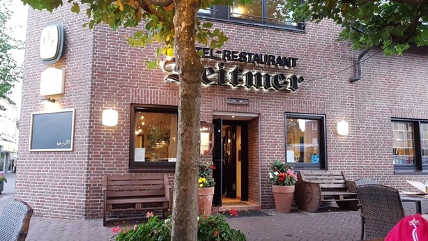 En byggnad av tegel med en skylt som visar texten ”RESTAURANG Seittimer”. Utomhus finns sittplatser med bänkar och planterade blommor.