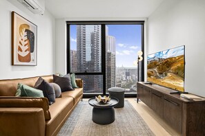 Un salon moderne avec un canapé en cuir, une table basse, un téléviseur et une grande fenêtre donnant sur la ville.