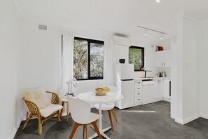 A modern kitchen with white cabinetry, a dining area with a round table and chairs, and a window with a view of trees. - Beachside 1-Bed Retreat with Parking Cronulla (Cronulla)