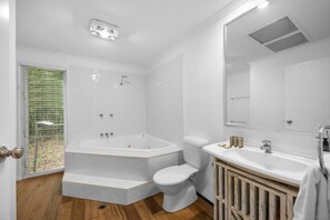 Et moderne badeværelse med et stort badekar, toilet og vask, med trægulv og et stort spejl.