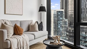 Un salon moderne avec un canapé gris, un lampadaire noir, une table basse ronde agrémentée de livres et d’un objet décoratif, et une grande fenêtre donnant sur la ville.