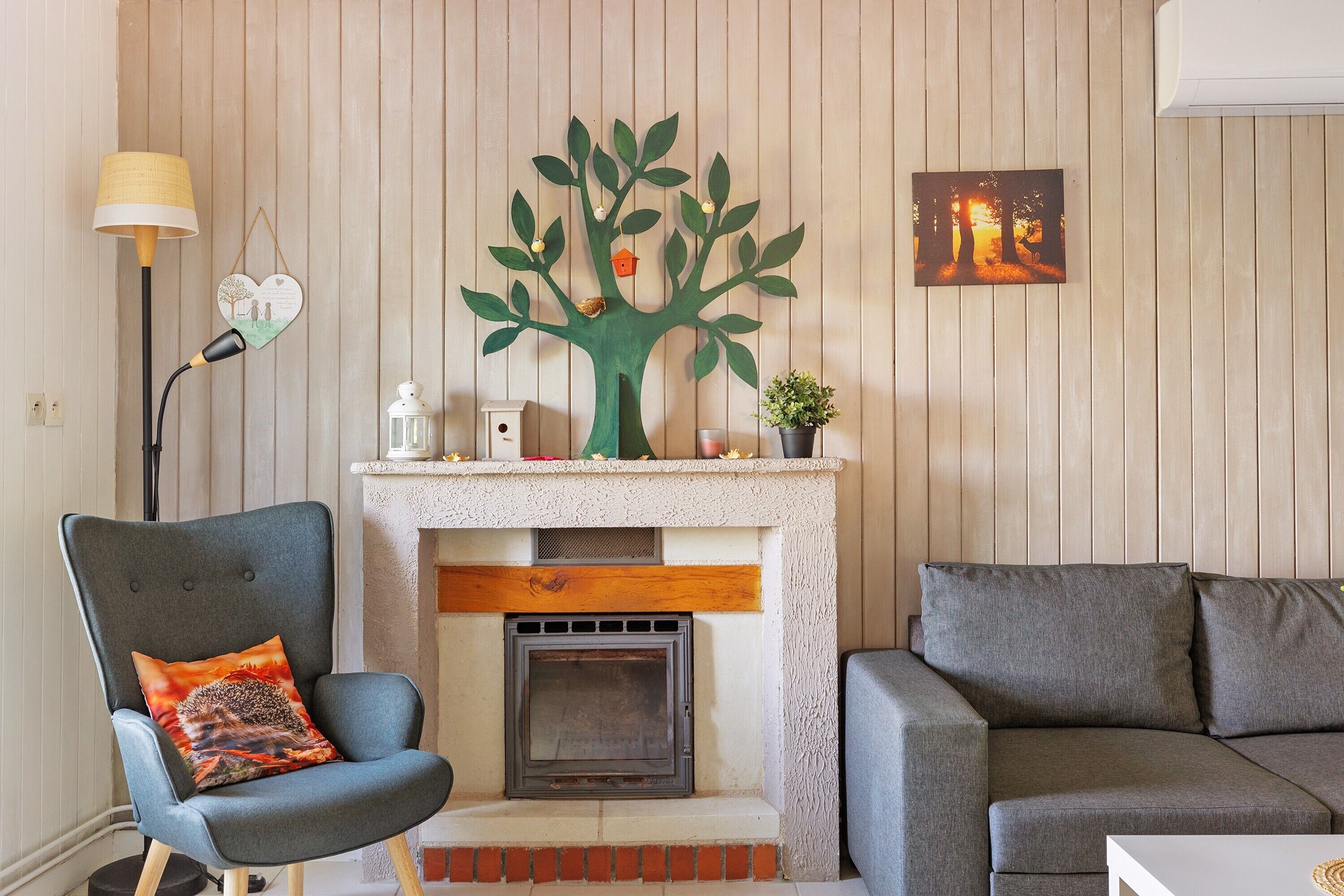 A living room with a fireplace, a blue armchair, a grey sofa, and a wall-mounted tree decoration.