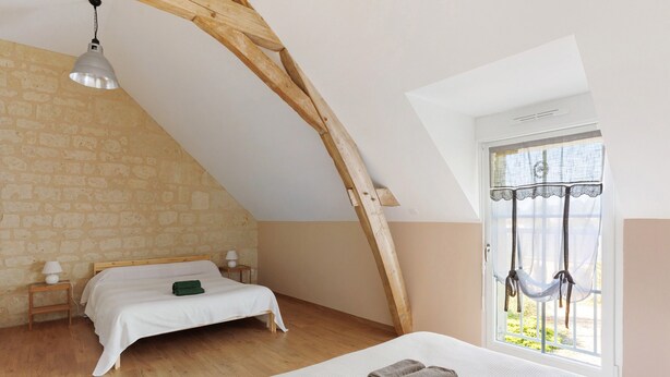 Ein Schlafzimmer mit schrägen Wänden, einer Hängematte am Fenster, einem Bett mit weißer Bettwäsche und einem hölzernen Nachttisch mit Lampe.
