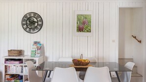 Ein Essbereich mit einem Glastisch, weißen Stühlen, einer Uhr, einem Bücherregal und einem gerahmten Bild an der Wand.