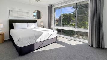 A modern bedroom with a large bed, a ceiling fan, a window with curtains, and a view of greenery outside.