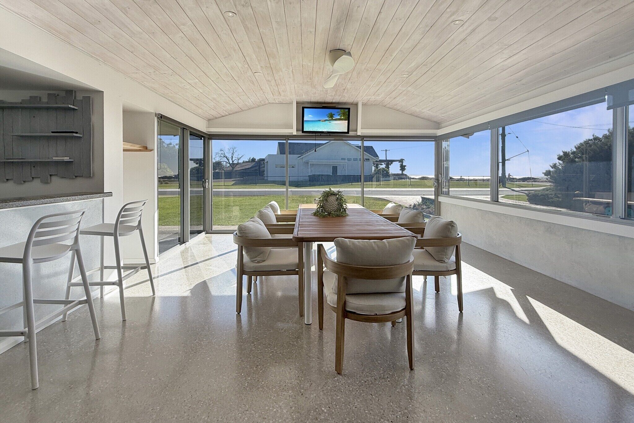 A modern dining area with a wooden table, white chairs, a TV on the ceiling, and large windows offering a view of the outdoors.