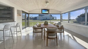 Uma sala de jantar moderna com uma mesa de madeira, cadeiras brancas, uma TV no teto e grandes janelas com vista para o exterior.