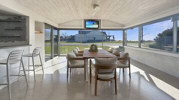 A modern dining area with a wooden table, white chairs, a TV on the ceiling, and large windows offering a view of the outdoors.