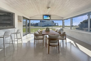 A modern dining area with a wooden table, white chairs, a TV on the ceiling, and large windows offering a view of the outdoors. - Comfortable Stay Near Busselton Beach (West Busselton)