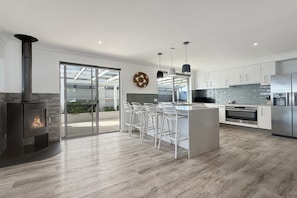 A modern kitchen with a central island, bar stools, a built-in stove, and a large glass door leading to an outdoor area. - Comfortable Stay Near Busselton Beach (West Busselton)