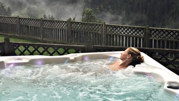 A hot tub with a person in it, surrounded by a wooden fence and a forested area.