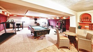 A pool table, exercise equipment, wicker chairs, and a bar area in a room with red walls and tiled flooring.