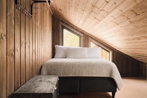 A loft bedroom with a sloped ceiling, wooden walls, a bed with a textured white bedspread, and a small wooden bench.