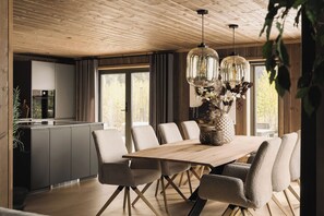 Een eetgedeelte met een houten tafel en stoelen, een keuken op de achtergrond en een groot raam met gordijnen.