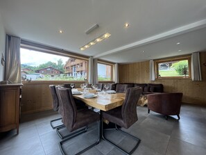 A dining area with a wooden table, cushioned chairs, and a view of the outside. - Chalet La Centaurée - by Care Concierge (Combloux)