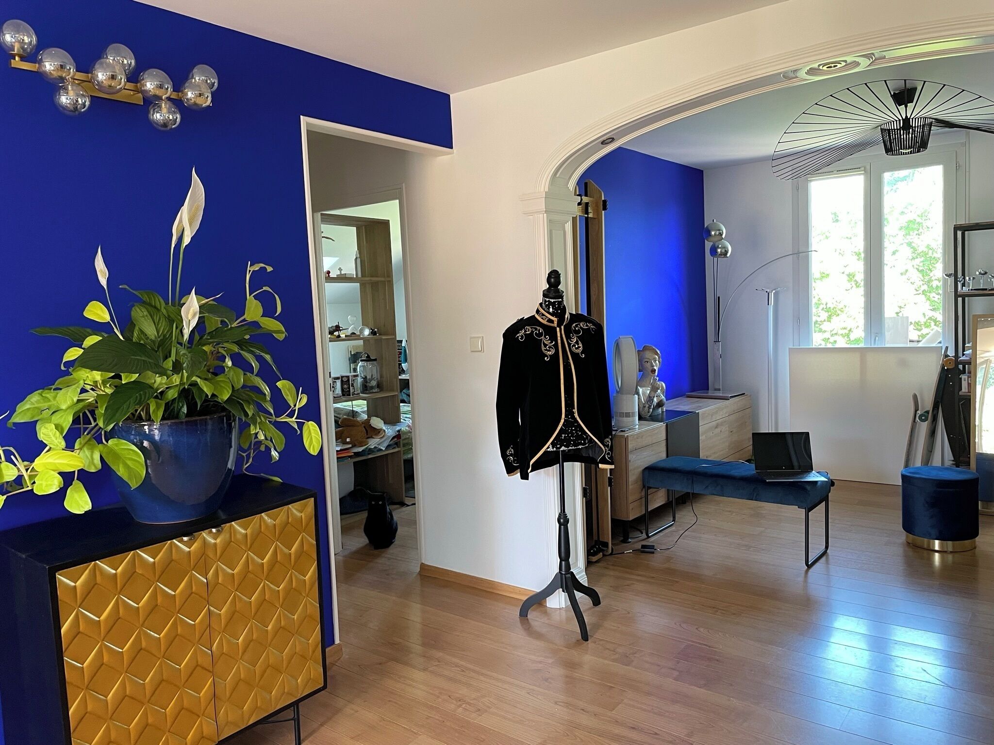 A room with a blue wall, a wooden floor, a plant, a mannequin, a desk with a laptop, and a large window.
