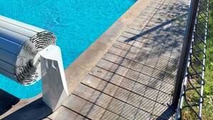 En rektangulär swimmingpool omgiven av ett trädäck, en byggnad i sten i bakgrunden och ett staket av metall.