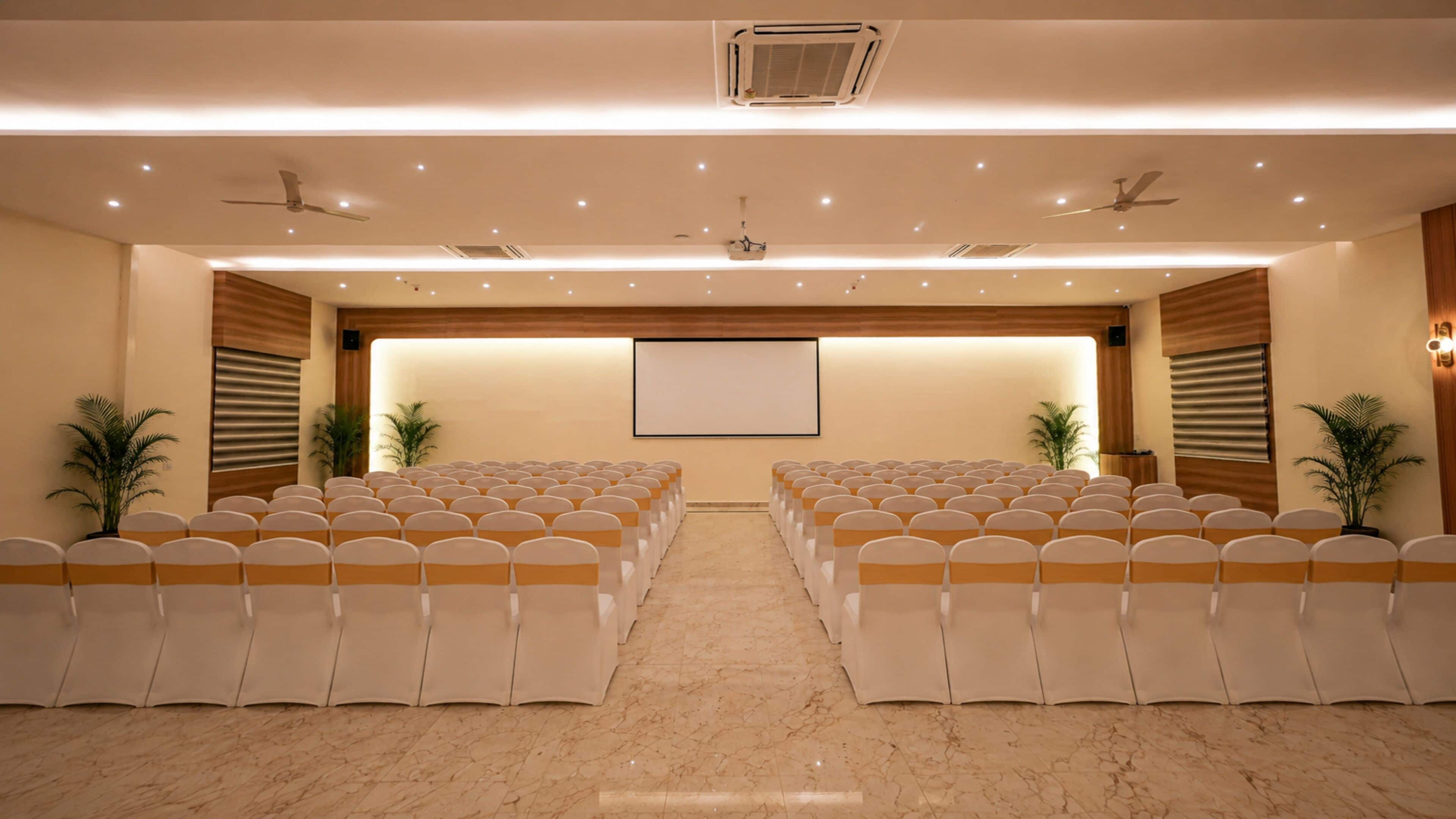 A conference room with rows of chairs, a projector screen, ceiling fans, and potted plants.
