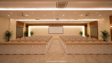 A conference room with rows of chairs, a projector screen, ceiling fans, and potted plants.