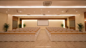 A conference room with rows of chairs, a projector screen, ceiling fans, and potted plants.
