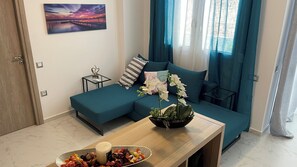 En moderne stue med en turkis sofa, et lille bord med skålsæt frugt og et stearinlys, samt et billede på væggen.
