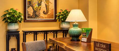 A reception desk with a chair, a lamp, two potted plants, and a framed artwork on the wall.