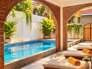 Piscina com borda azulejada, rodeada por arcos de tijolo, plantas em vasos e espreguiçadeiras com toalhas.