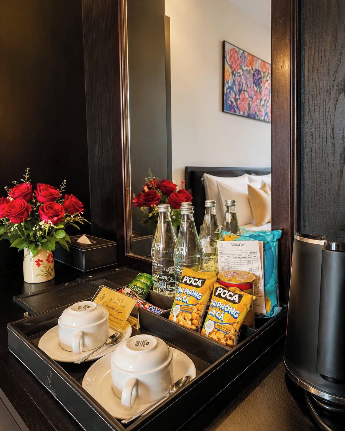 Habitación de hotel con mesita de noche, espejo, cama con almohadas, un cuadro en la pared y una bandeja con tazas de café, botellas, aperitivos y una planta.