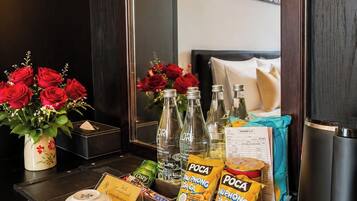 Habitación de hotel con mesita de noche, espejo, cama con almohadas, un cuadro en la pared y una bandeja con tazas de café, botellas, aperitivos y una planta.