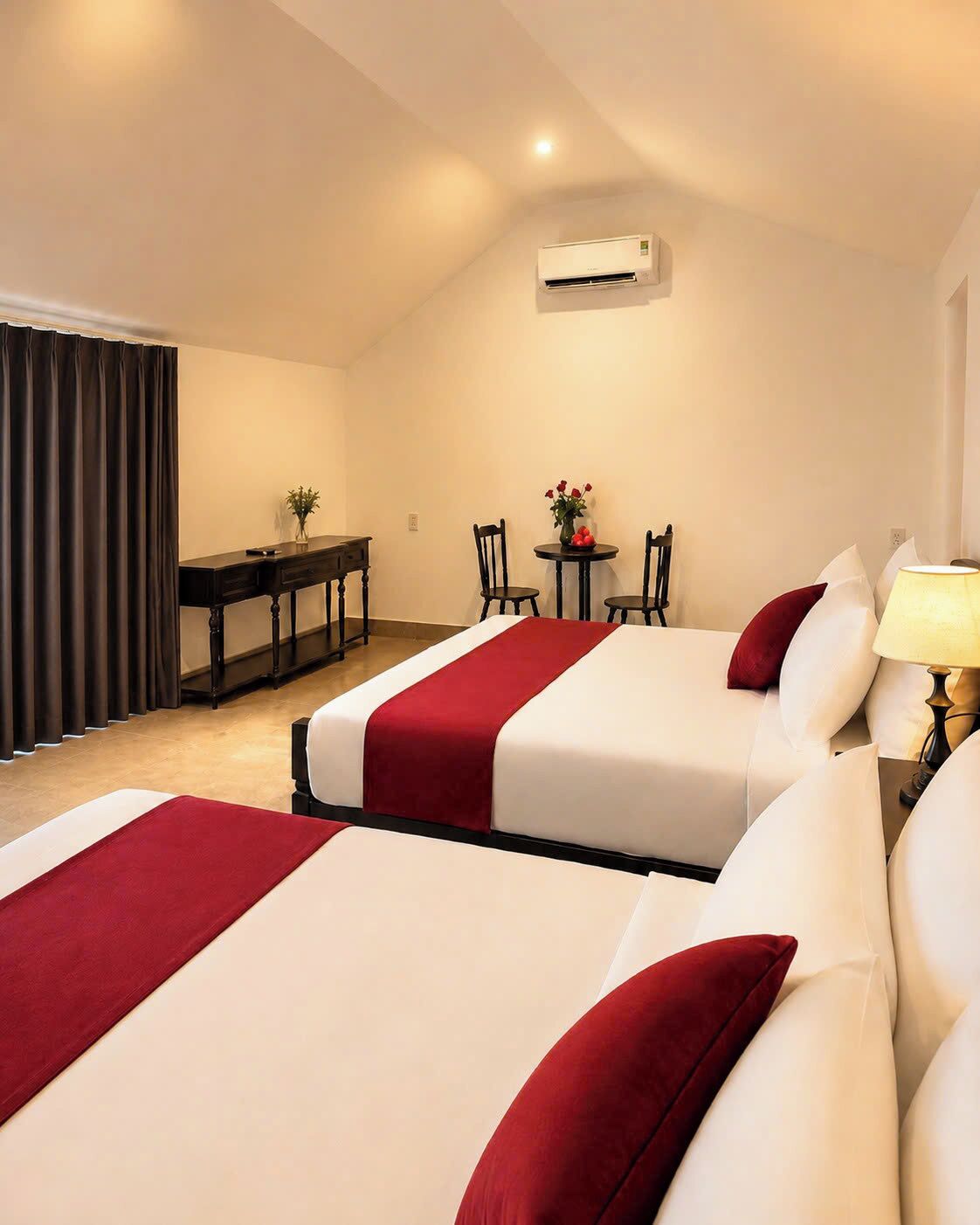 A hotel room with two beds, a desk, a chair, a table with flowers, and an air conditioning unit.