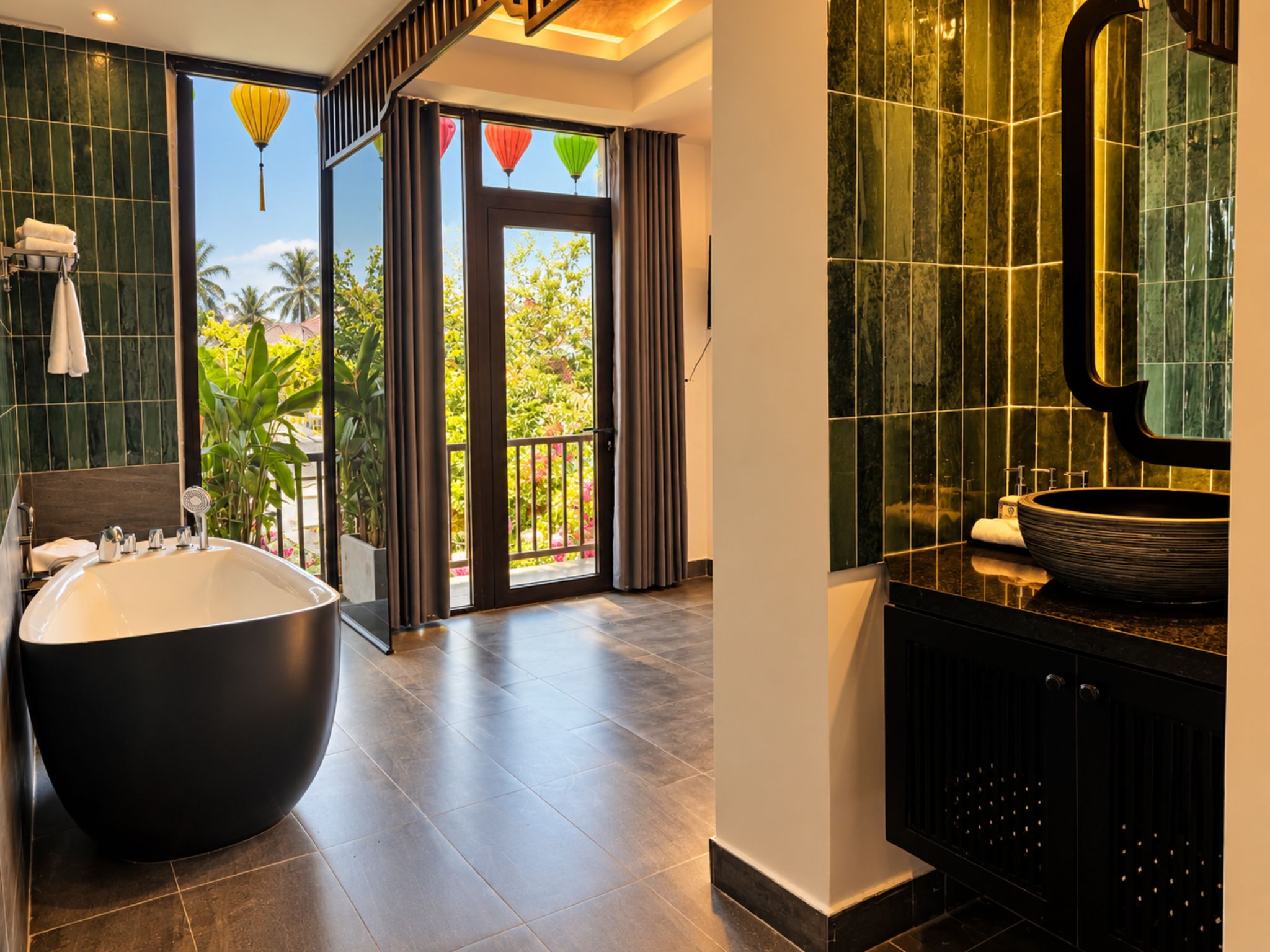 A modern bathroom with a large bathtub, a round mirror, and a view of a balcony with plants.