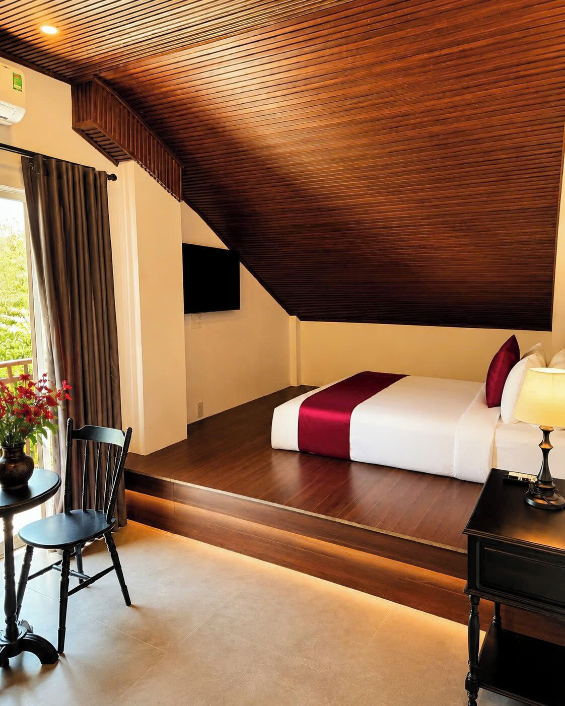 Habitación de hotel con techo de madera, cama con colcha roja y blanca, mesita de noche con lámpara, una silla y una mesa con un jarrón de flores.