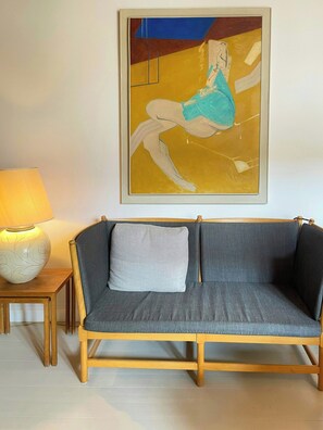 Ein Zimmer mit grauem Sofa, einem kleinen Tisch mit Lampe und einem gerahmten abstrakten Gemälde an der Wand.