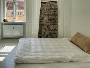 Ein Schlafzimmer mit einem Bett, einem Heizkörper, einem Fenster mit Blick auf Gebäude und einem gemusterten Vorhang.