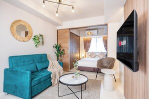 A modern hotel room with a teal sofa, a round coffee table, a TV, and a bed with a wooden wardrobe.