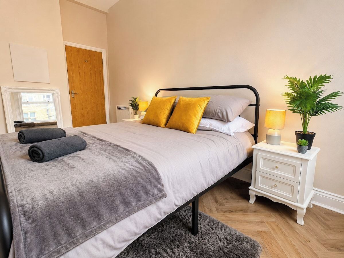 Un lit avec une couverture grise, deux oreillers, une table de chevet avec une lampe et une plante en pot.