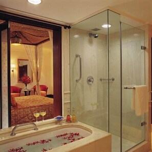A bathroom with a glass shower enclosure, a bathtub with rose petals, and a mirror reflecting a room with a bed and a table. - Itc Rajputana A Luxury Collection Hotel (Jaipur)