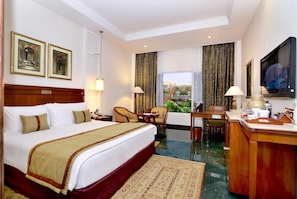 A hotel room with a large bed, a desk, a chair, a television, and a view of the city through the window. - Itc Rajputana A Luxury Collection Hotel (Jaipur)