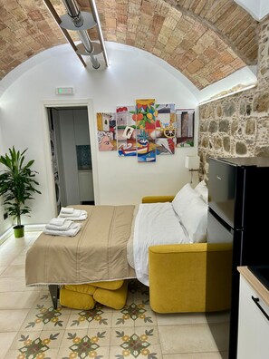 Habitación de hotel con cama, sofá, zona de comedor, planta en maceta y una pared decorada con obras de arte.