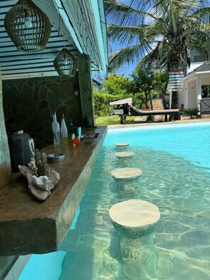 Un bar junto a la piscina con escalones de piedra que bajan al agua, una palmera y un muro de piedra.