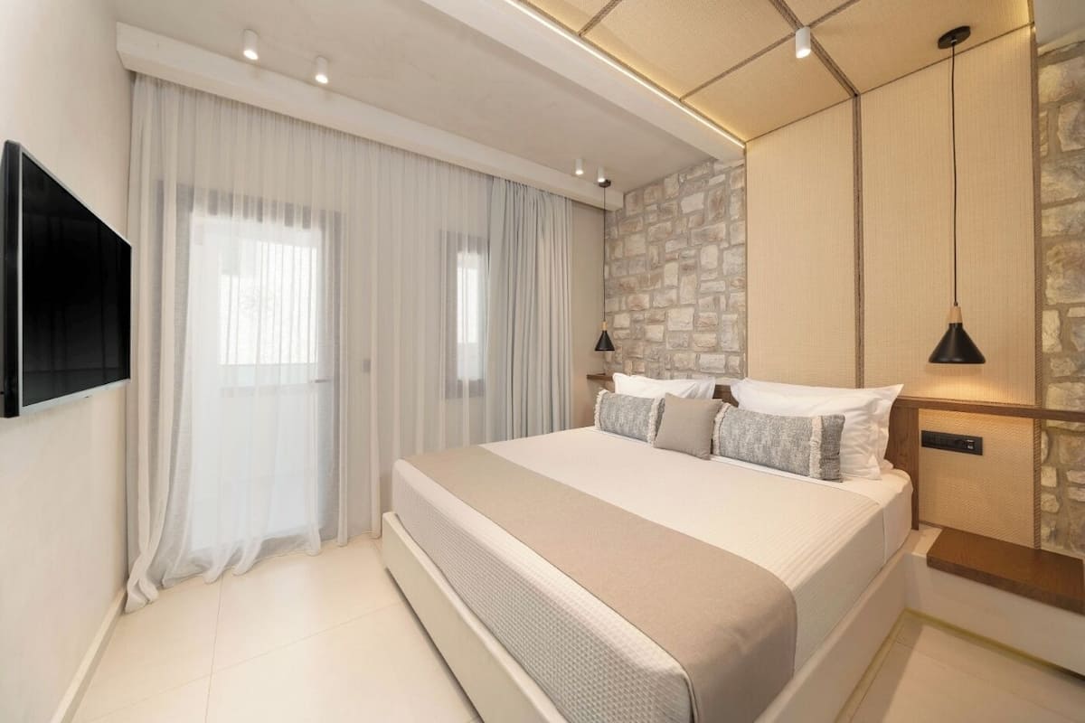Ein modernes Schlafzimmer mit einem großen Bett, einer Wand aus Stein, einem angebrachten Fernseher und einem Fenster mit transparenten Vorhängen.