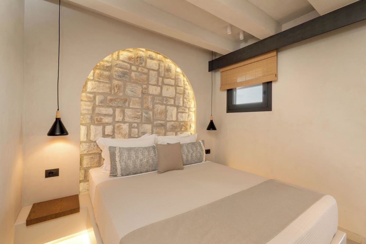 Ein Schlafzimmer mit einer Steinkuppel, einem Bett mit weißer Bettwäsche, zwei Pendelleuchten und einem Fenster mit Bambusrollvorhang.