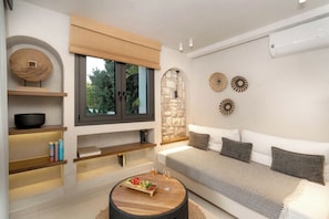 A modern living room with a sofa, a coffee table with fruit, a bookshelf, and a window with a view. - The Sand Suites (Agios Nikolaos)
