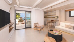 A modern living room with a TV, sofa, dining table, and a view of the sea. - The Sand Suites (Agios Nikolaos)