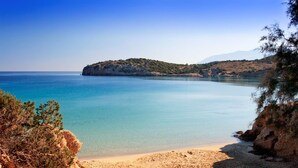 A coastal landscape with a sandy beach, clear blue water, and a rocky hill in the background. - The Sand Suites (Agios Nikolaos)